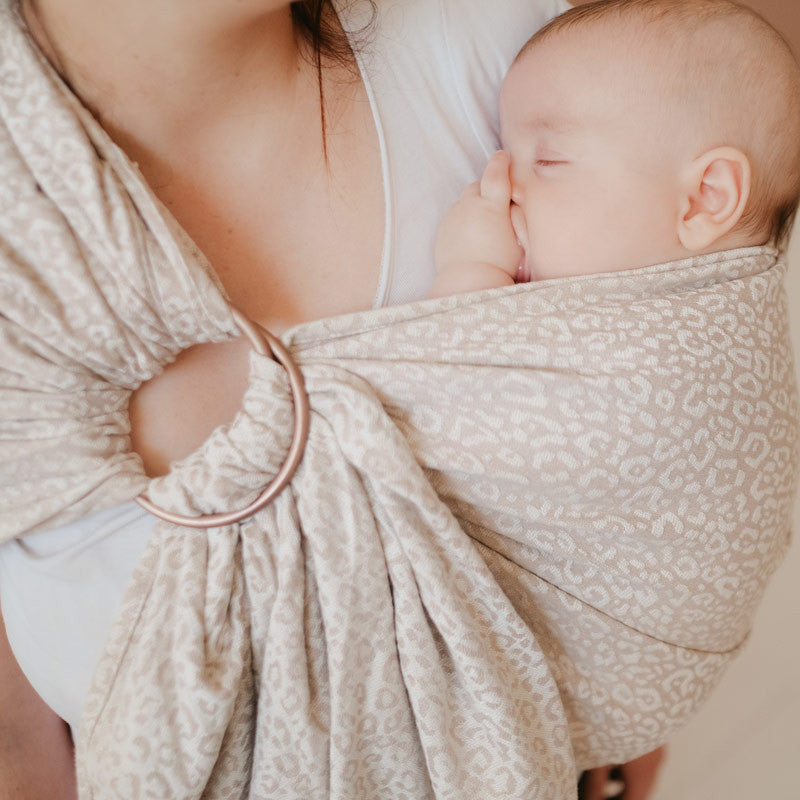 Mãe usando um sling de argolas Neobulle JACQUARD Léopard Beige, com o bebé a dormir encostado ao peito.