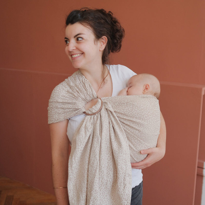 Mulher sorridente usando o sling de argolas Neobulle JACQUARD Léopard Beige com um bebé, demonstrando a facilidade de uso e o ajuste perfeito.