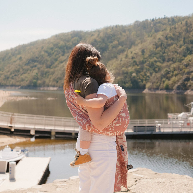 Mãe com Sling de Argolas Neobulle SEA CELL azalée rose, posicionada ao ar livre com paisagem de lago ao fundo.