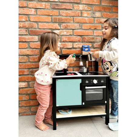 Duas crianças a brincar com a cozinha industrial Small Foot, uma a usar o fogão e a outra a encher o tacho, num ambiente com tijolo aparente.