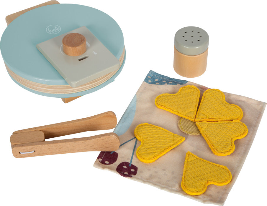 Vista geral dos acessórios do conjunto de waffles de brincar: máquina fechada, waffles de tecido, pinça de madeira e doseador de açúcar sobre fundo branco.