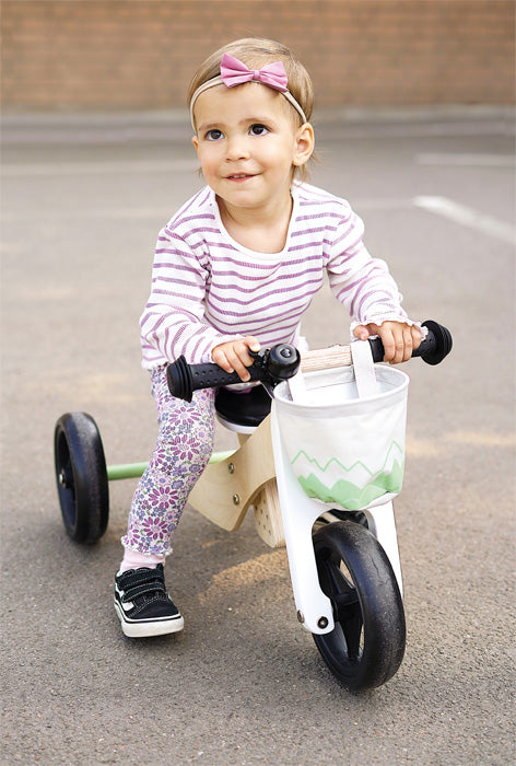 Criança pequena sorridente a andar num triciclo de treino 2 em 1 Small Foot, com cesto branco e verde, ao ar livre.