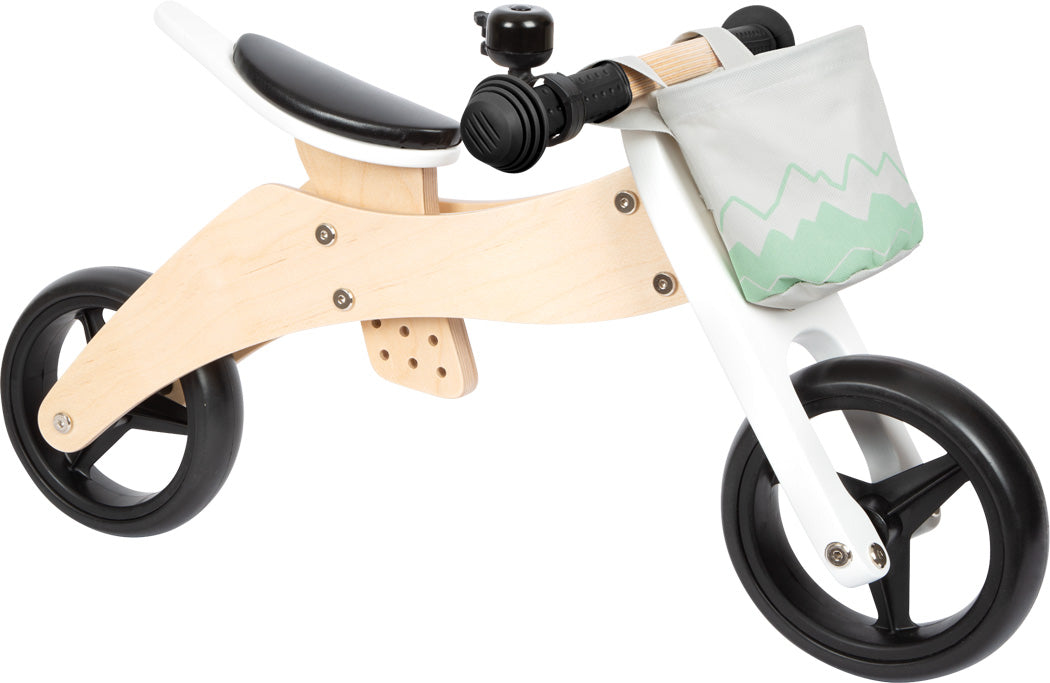 Triciclo de treino Small Foot com foco na parte frontal, exibindo o guiador com punhos pretos, campainha e cesto de tecido cinza com padrão de montanha verde.
