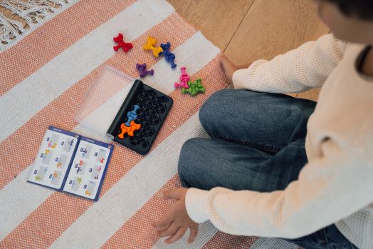 Vista superior de uma criança sentada no chão a jogar o IQ Waves, com o tabuleiro e as peças espalhadas ao lado do livro de desafios.