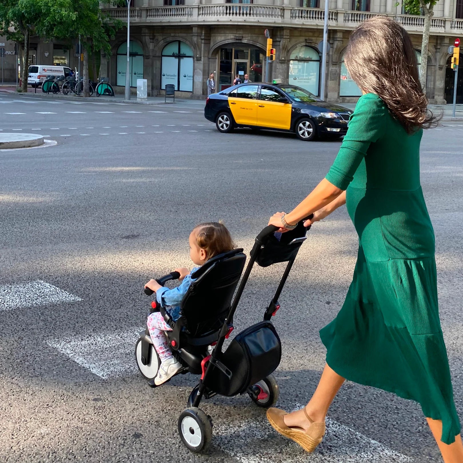 Mãe grávida empurra o SmartTrike STR7, usado como carrinho de passeio, com um bebé sentado, a atravessar uma passadeira urbana.