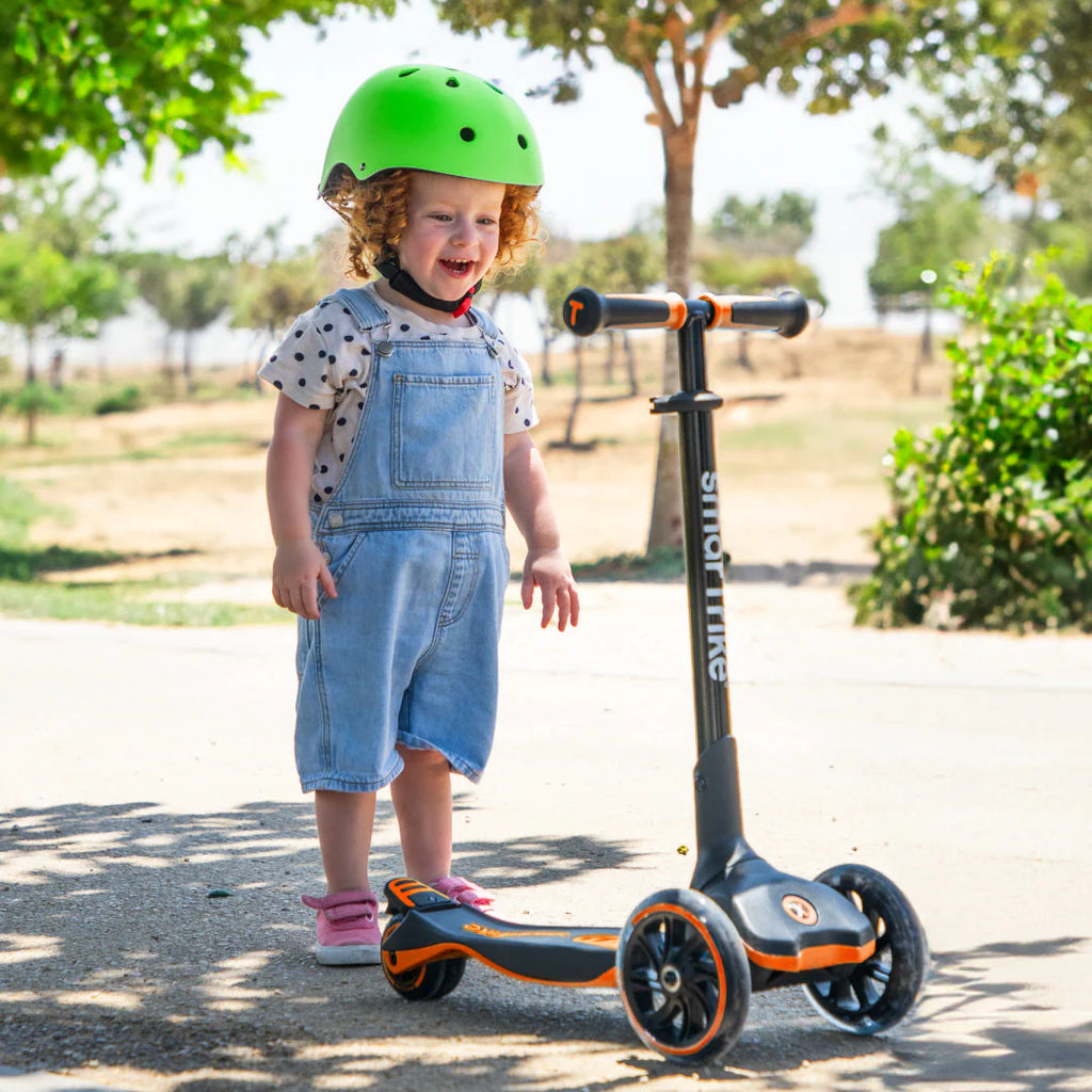 Criança pequena com capacete verde e jardineiras de ganga sorri ao lado da trotinete Smartrike Xtend Ride-on, modo inicial.