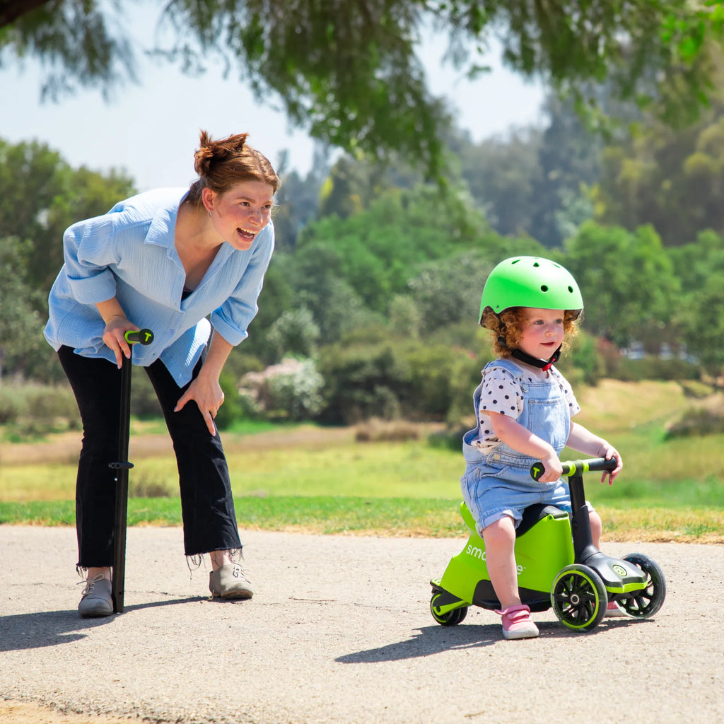 Criança pequena com capacete verde a usar a Smartrike Xtend no modo "ride-on" (com assento), supervisionada por uma adulta num parque.