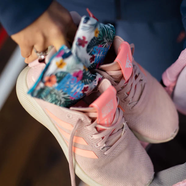 Mãos inserem dois sacos SmellWell Active com padrão floral tropical em ténis desportivos cor-de-rosa, demonstrando o uso para absorver humidade e odor.