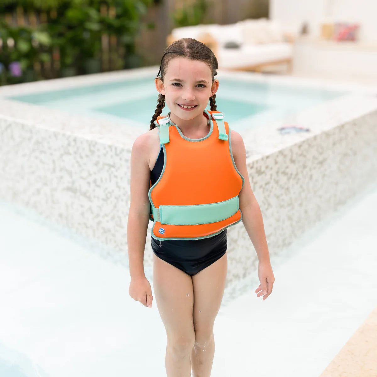 Menina sorridente a usar o colete natação Splash About Go Splash Adjustable Swim Vest Laranja e Verde ao lado da piscina.