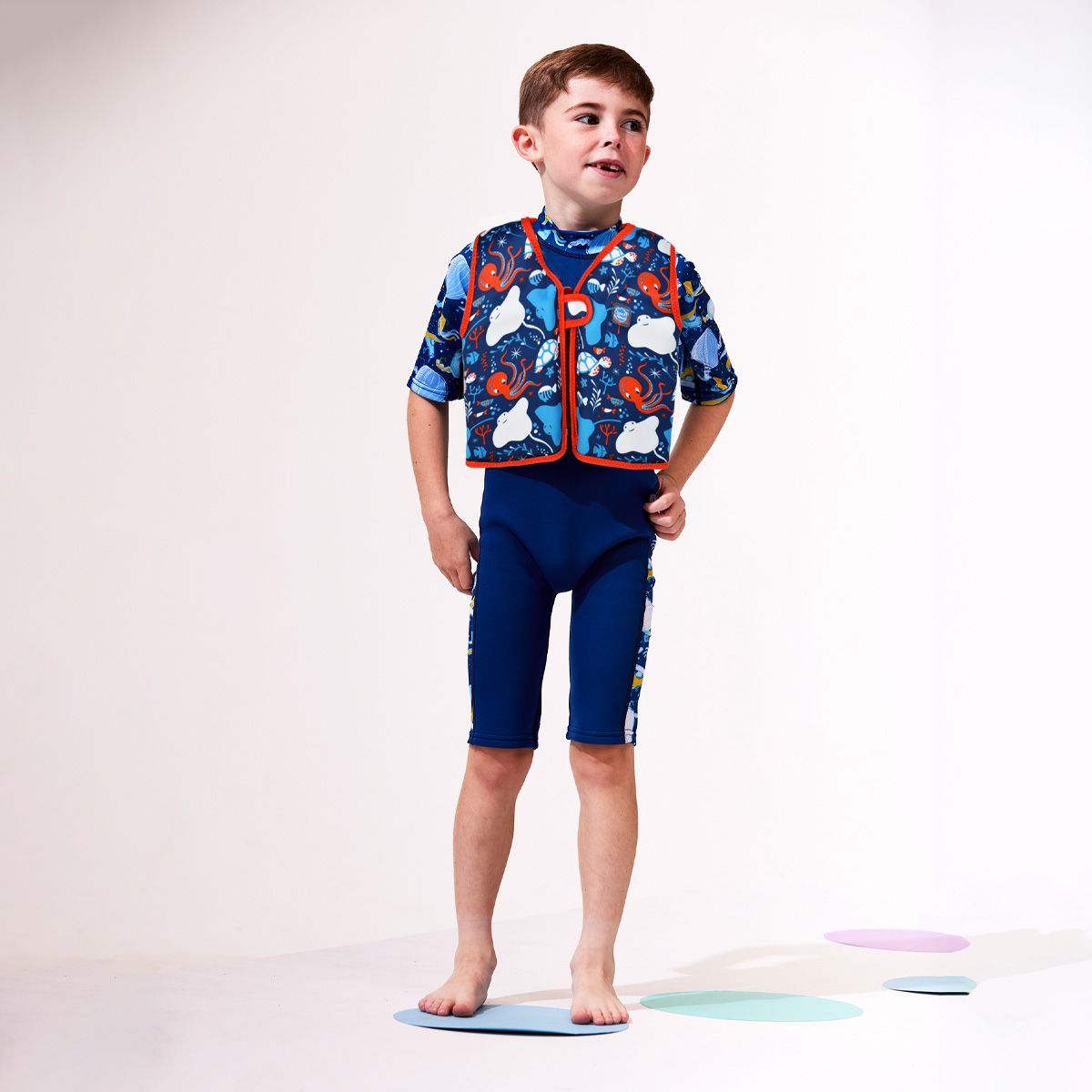 Criança a usar o colete natação Splash About Float Jacket com padrão marinho, pronto para aprender a nadar.
