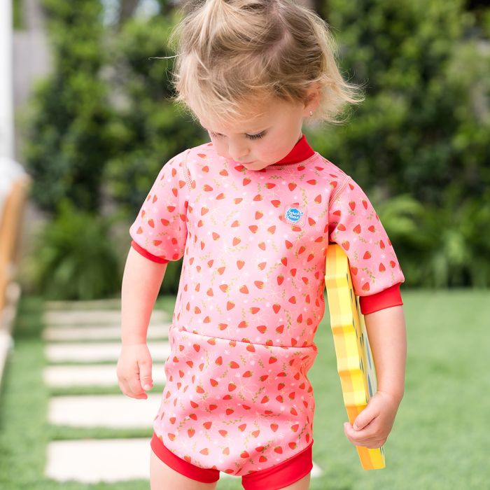 Criança pequena com o fato de banho térmico de morangos, olhando para baixo enquanto segura um acessório de natação amarelo, no exterior.