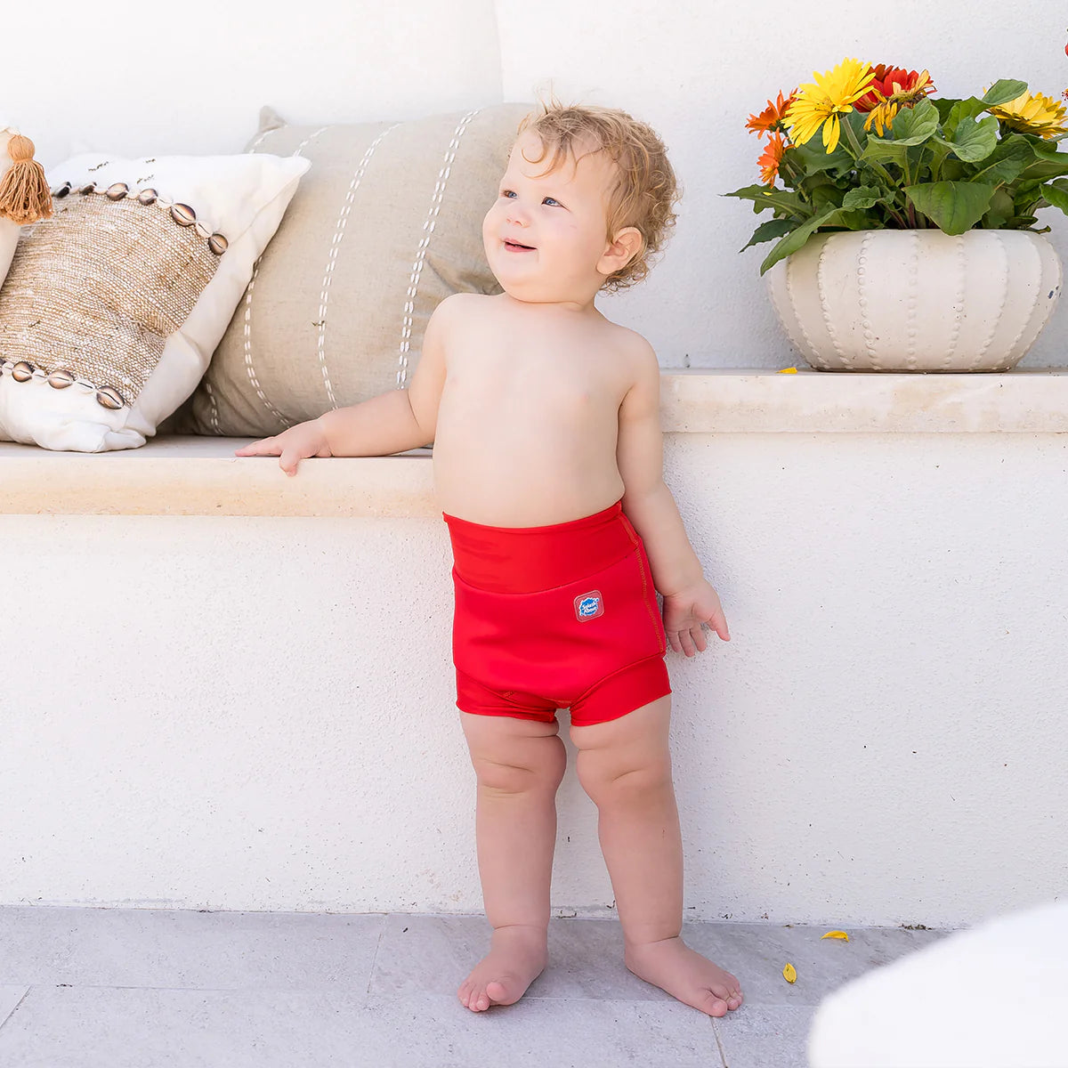 Bebé com fralda de natação Happy Nappy vermelha, a olhar para cima com um sorriso, em ambiente exterior ensolarado.