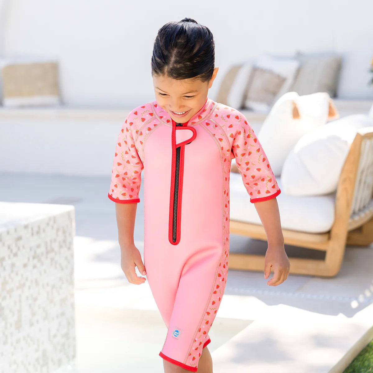 Criança a caminhar num pátio ensolarado, sorrindo, vestindo o fato de praia/piscina térmico rosa com padrão de morangos.