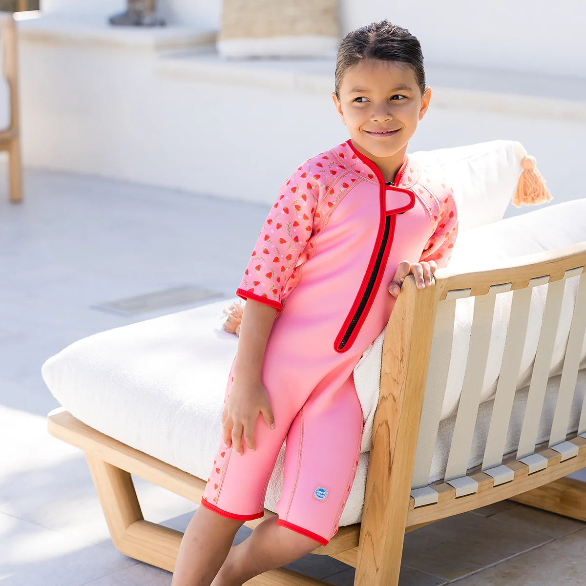 Criança sorridente encostada a uma espreguiçadeira branca, vestindo o fato de banho térmico de manga curta com padrão de morangos.