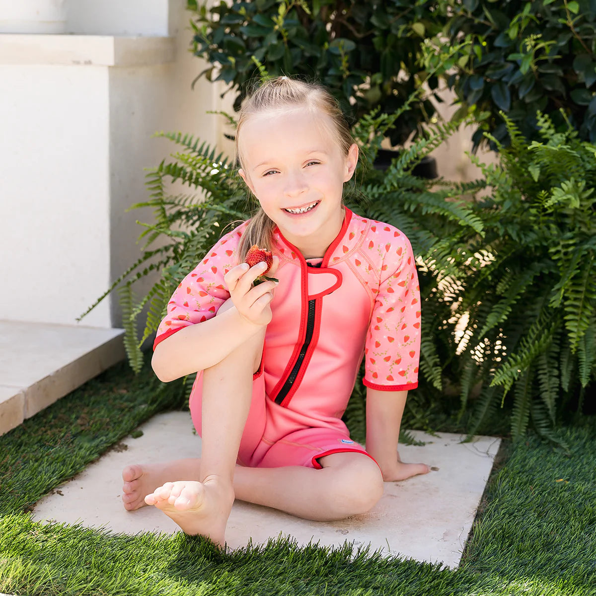 Criança sorridente sentada num pátio com relva, vestindo o fato de banho térmico de morangos e segurando um morango.