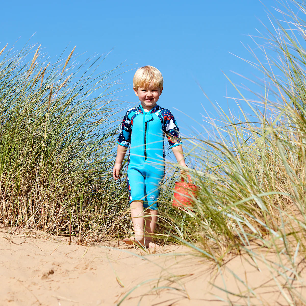 Menino loiro sorridente a usar o fato de neoprene azul 'Under the Sea' na praia, caminhando entre dunas de relva alta sob o sol.