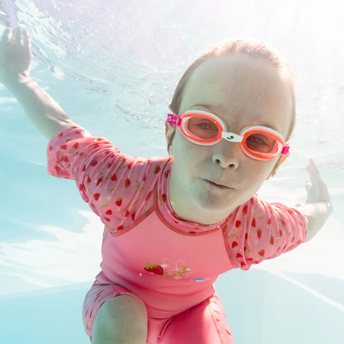 Criança a nadar debaixo de água com o fato térmico de morangos, usando óculos cor-de-rosa, com luz solar a penetrar na água.
