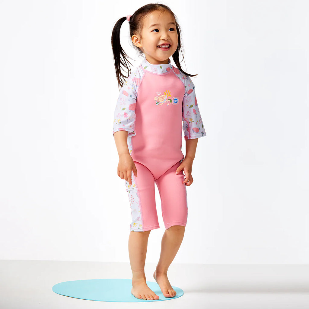 Criança sorridente a usar o fato de banho térmico Splash About UV Sun & Sea, ideal para praia ou piscina, em pose de pé.