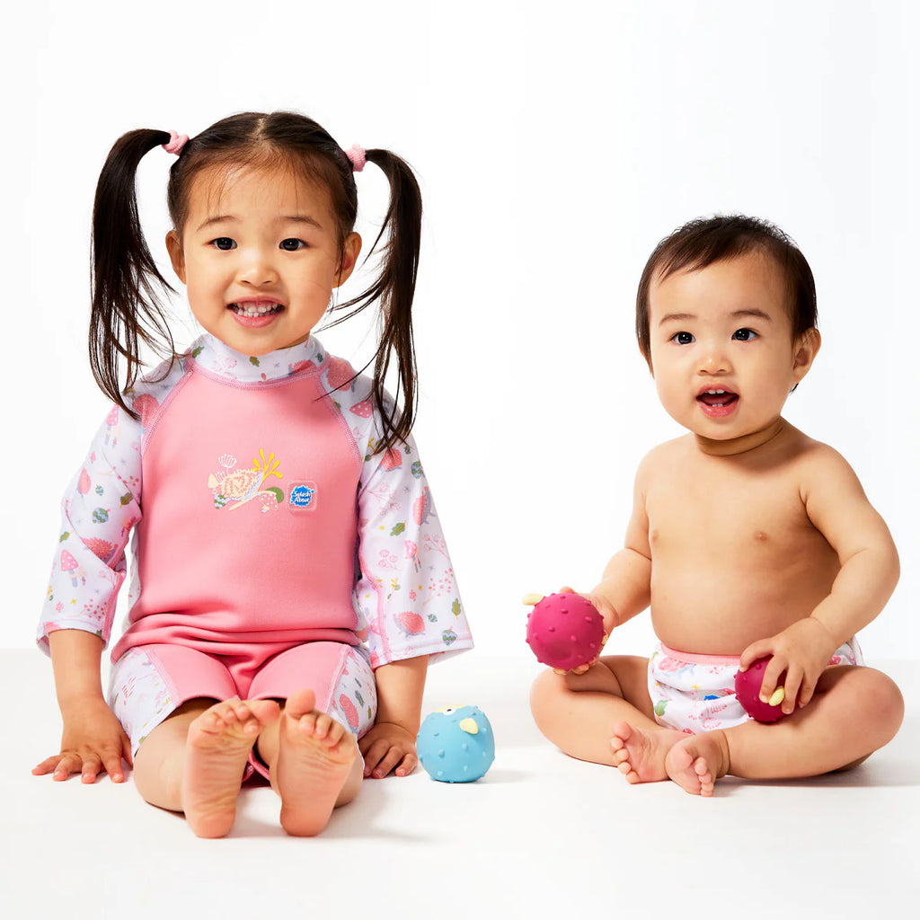 Duas crianças, uma mais velha a usar o fato térmico e um bebé, a brincar em chão branco com brinquedos.