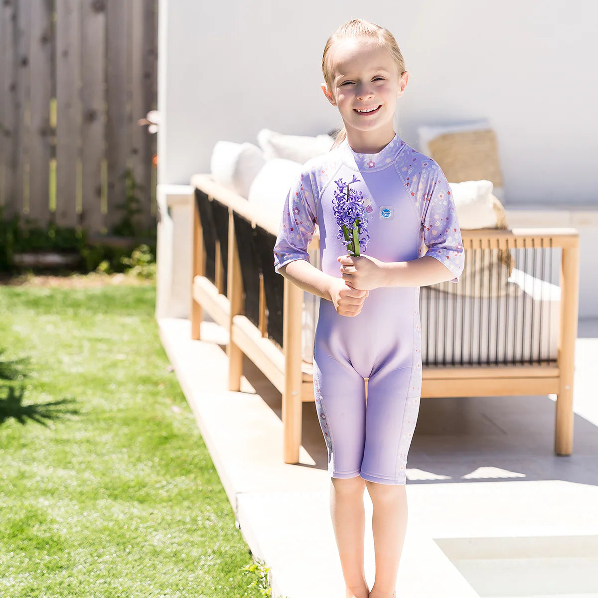 Criança sorridente com cabelo loiro, vestindo o fato de banho térmico lilás, segurando flores num pátio exterior com relva e mobiliário de exterior.