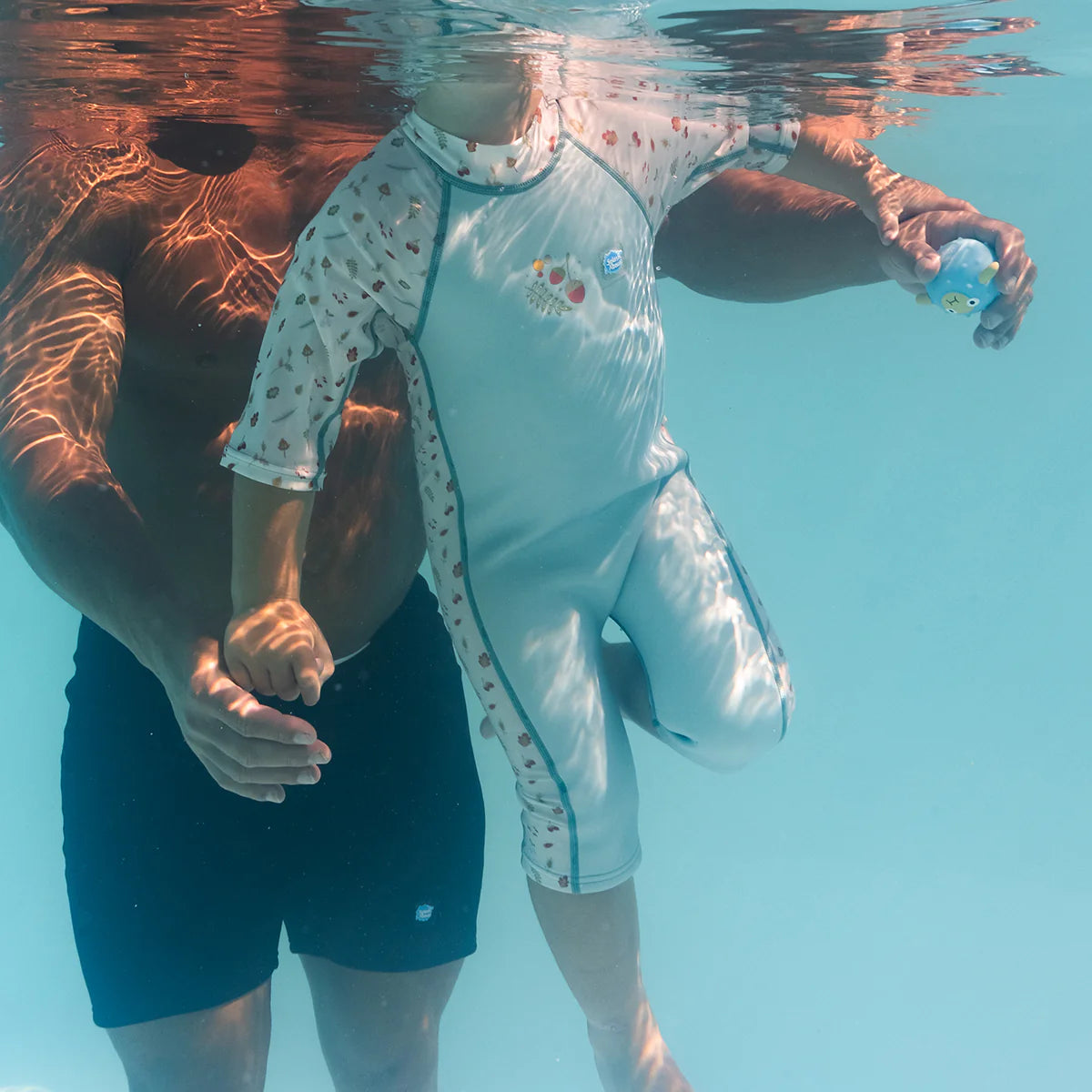 Criança a usar o fato de mergulho UV Sun & Sea a flutuar debaixo de água com um adulto a segurá-la na piscina.