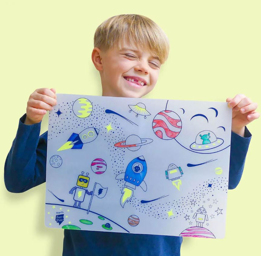 Criança sorridente a exibir o tapete de silicone SuperPetit totalmente colorido com desenhos de astronautas, planetas e foguetões.