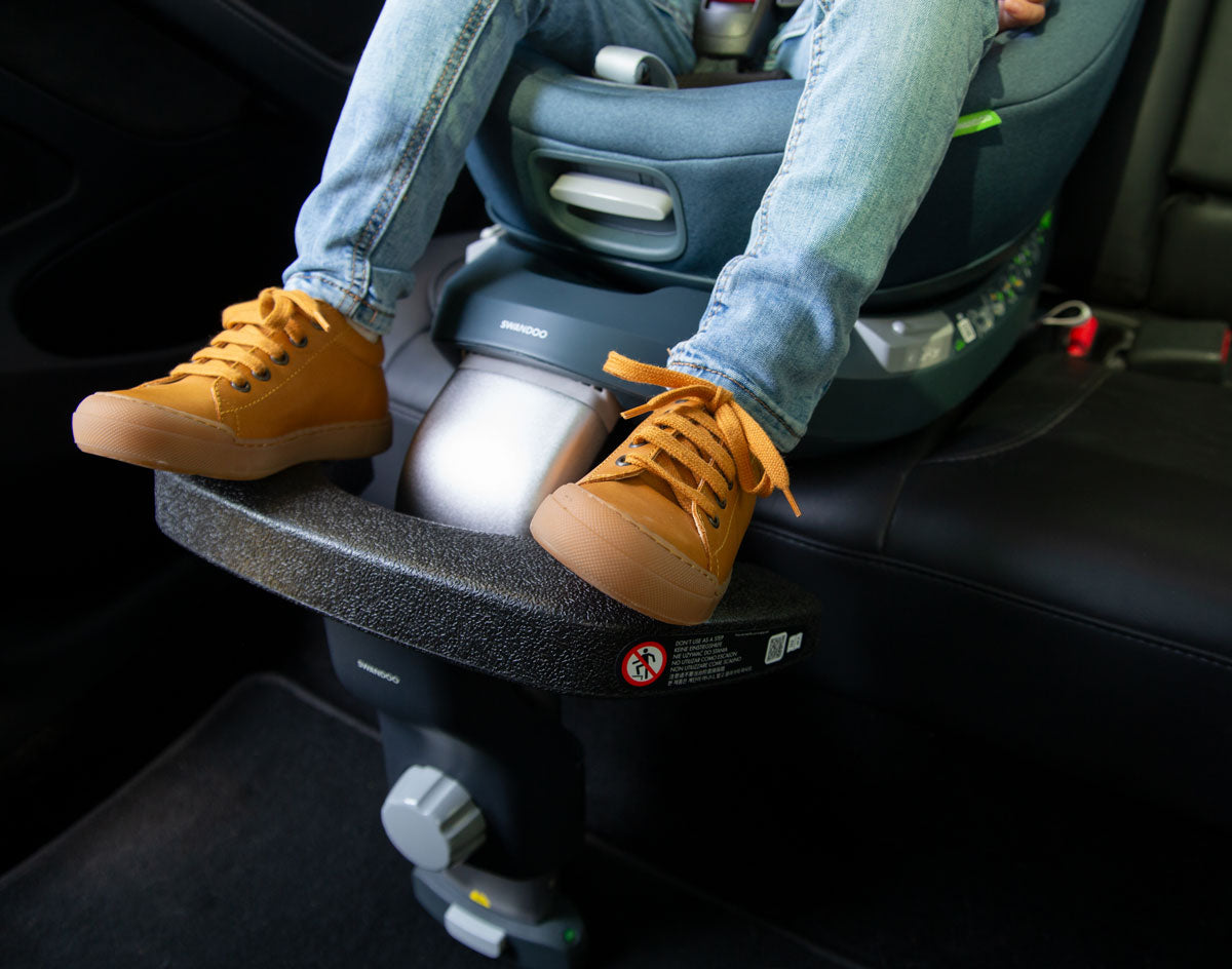 Criança com sapatos amarelos sentada num assento auto, a usar o apoio universal Swandoo, que sustenta confortavelmente os seus pés.