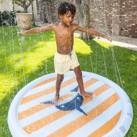 Criança a brincar num tapete de esguichos inflável Swim Essentials, com design de tubarão, num jardim ensolarado.