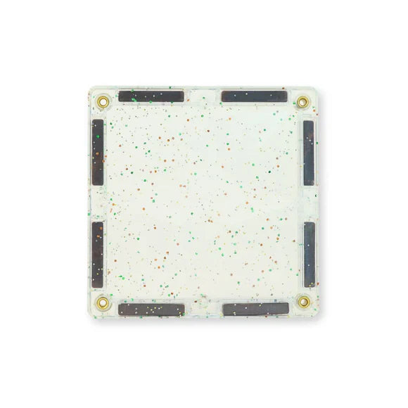 Vista de perto de uma telha magnética quadrada transparente com salpicos de glitter verde e castanho, mostrando os ímanes laterais.