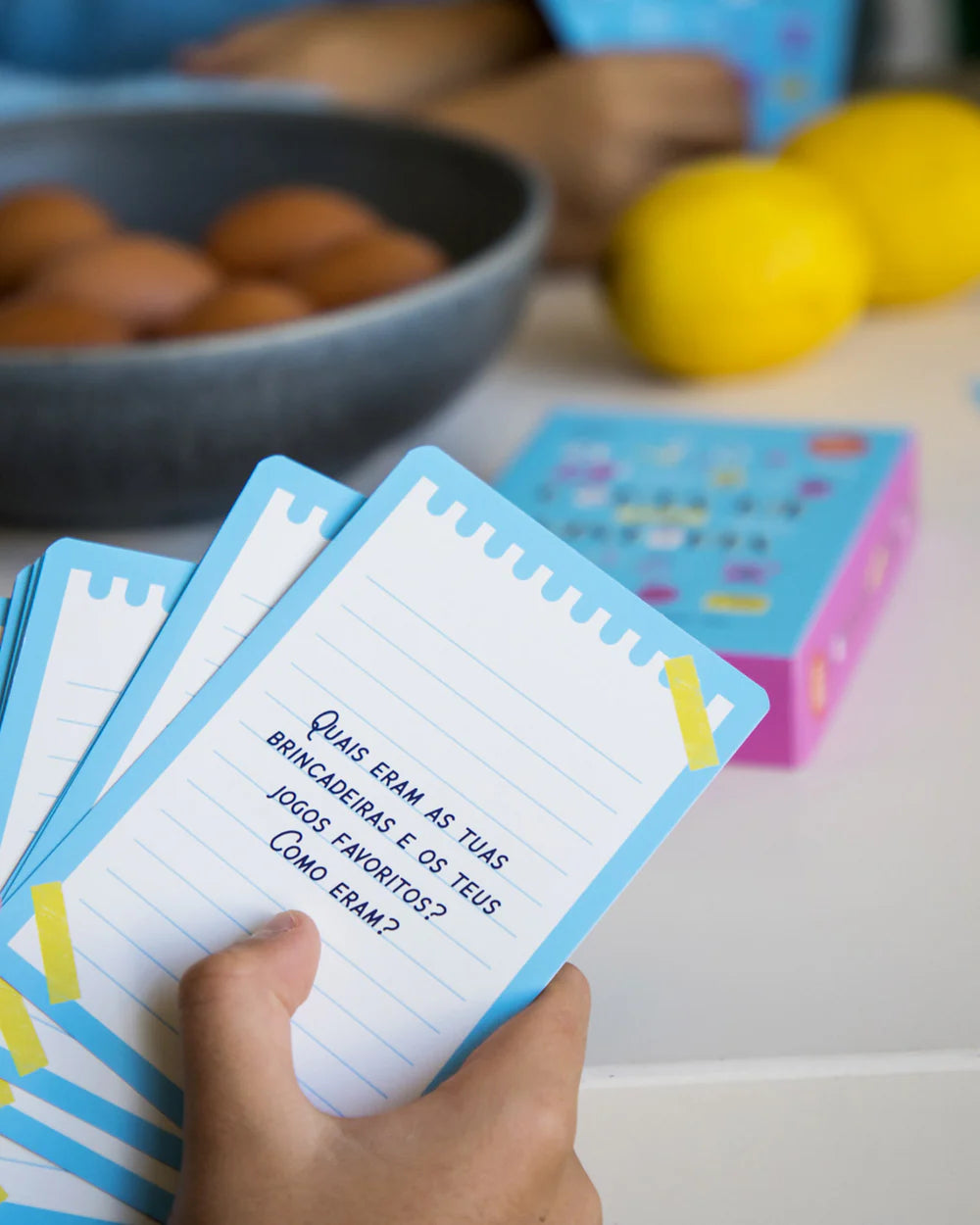 Mão segurando cartas do jogo "Conta-me a Tua História", mostrando perguntas sobre brincadeiras e jogos de infância.