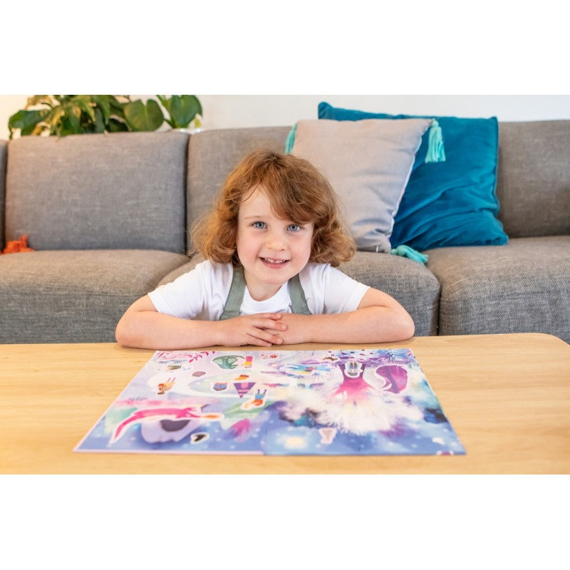 Criança com cabelo encaracolado a olhar diretamente para a câmara, com o livro de autocolantes aberto do Mundo Mágico na mesa à sua frente.