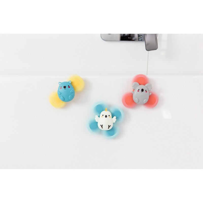 Os três brinquedos giratórios sensoriais fixados numa superfície branca, possivelmente uma banheira, em uso durante a hora do banho.