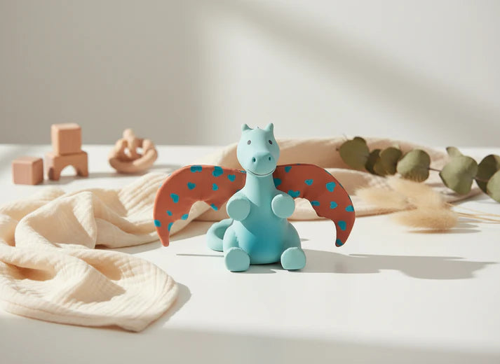 Brinquedo chocalho dragão Tikiri num cenário de quarto de bebé com luz natural, sobre um tecido bege e rodeado de elementos decorativos de madeira.