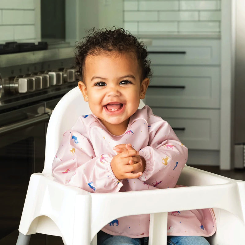 Bebé sorridente com cabelo encaracolado a usar o babete impermeável de unicórnios, sentado numa cadeira alta na cozinha.