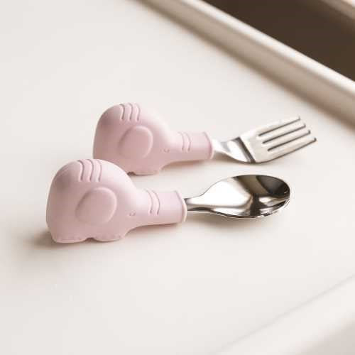 Detalhe dos talheres de aprendizagem Tiny Twinkle (colher e garfo) com cabos de silicone rosa em forma de elefante, pousados numa superfície branca.