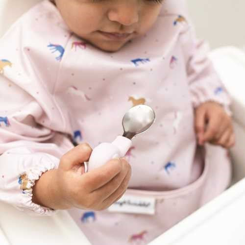 Bebé a usar um babete de cavalos, segurando com firmeza a colher de aprendizagem Tiny Twinkle com cabo de elefante rosa.