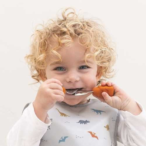 Criança pequena de cabelo encaracolado a usar a colher Tiny Twinkle com cabo de urso laranja para comer, vestindo um babete de dinossauros.