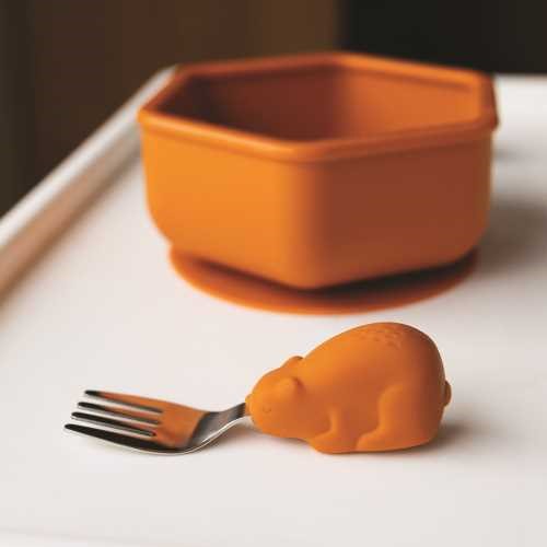 Garfo Tiny Twinkle com cabo de urso laranja ao lado de uma taça de silicone hexagonal laranja numa bandeja de bebé.