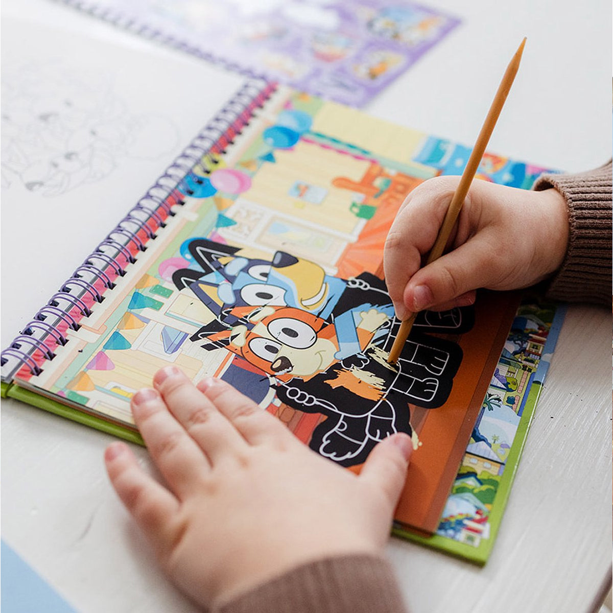 Mãos de uma criança a usar a caneta de madeira para raspar uma das placas pretas do livro Bluey, revelando cores por baixo, focando na atividade prática.