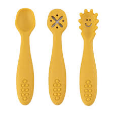 Três colheres de silicone amarelo mostarda lado a lado sobre fundo branco: uma colher normal, uma com orifícios em forma de X e uma com ponta texturada e um rosto sorridente.