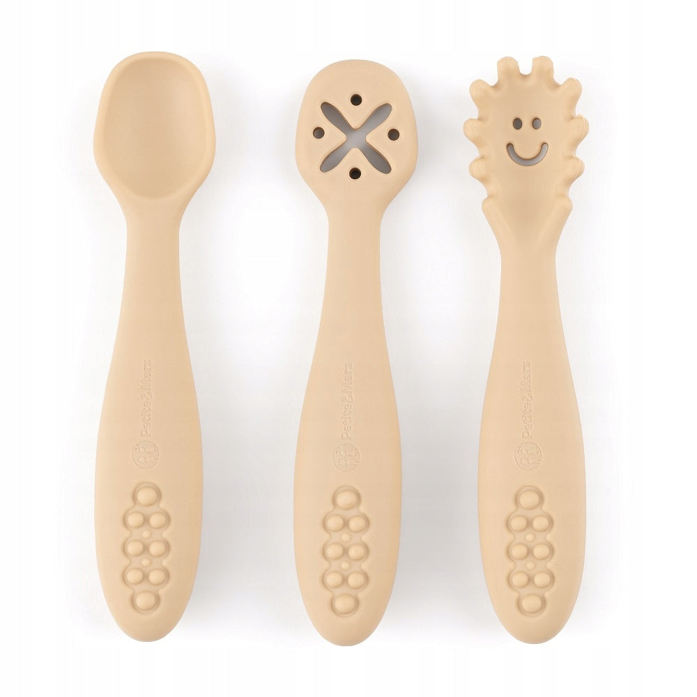 Três colheres de silicone bege lado a lado sobre fundo branco: uma colher normal, uma com orifícios em forma de X e uma com ponta texturada e um rosto sorridente.