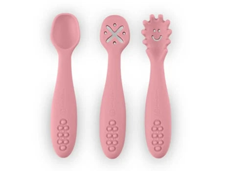 Três colheres de silicone cor-de-rosa lado a lado sobre fundo branco: uma colher normal, uma com orifícios em forma de X e uma com ponta texturada e um rosto sorridente.