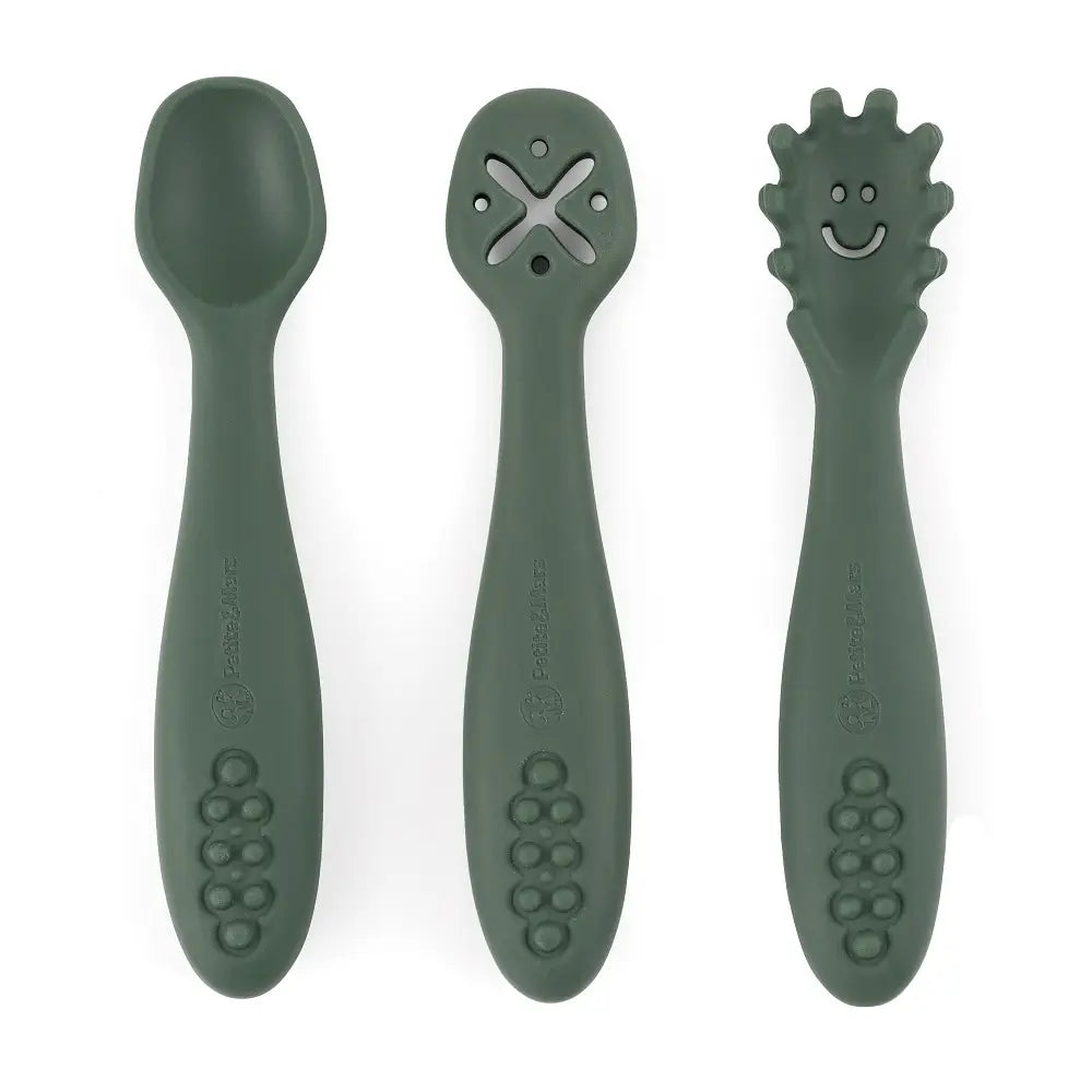 Três colheres de silicone verde-escura lado a lado sobre fundo branco: uma colher normal, uma com orifícios em forma de X e uma com ponta texturada e um rosto sorridente.