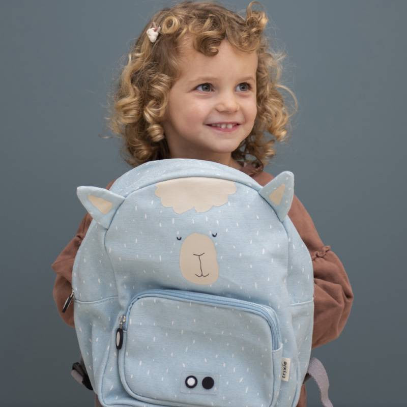 Criança de cabelo encaracolado a sorrir, segurando a mochila Trixie Alpaca azul clara à frente do corpo.