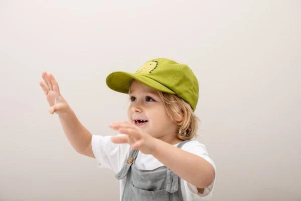 Criança pequena com cabelo loiro a usar o boné de dinossauro verde-lima, com os braços levantados, num ambiente claro.