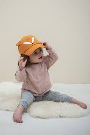 Criança pequena a usar o boné de pala Trixie raposa, ajustando-o, sentada em peles sintéticas brancas.