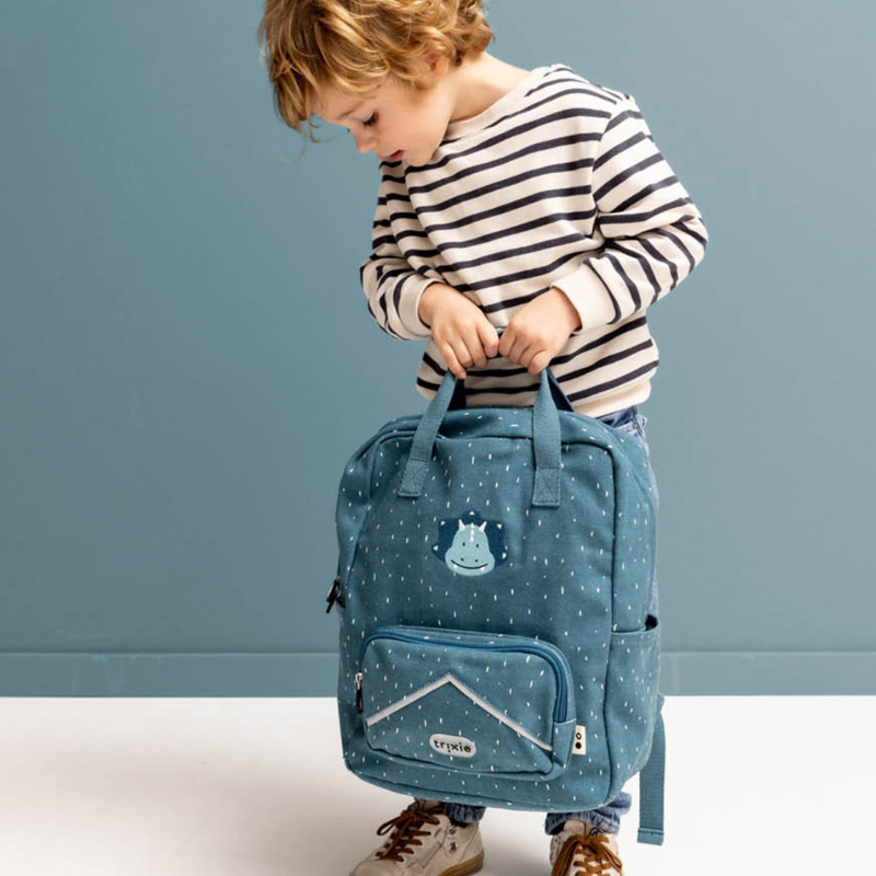 Criança com cabelo loiro a segurar a mochila escolar Trixie Triceratops azul-petróleo com padrão de salpicos, vista frontal.