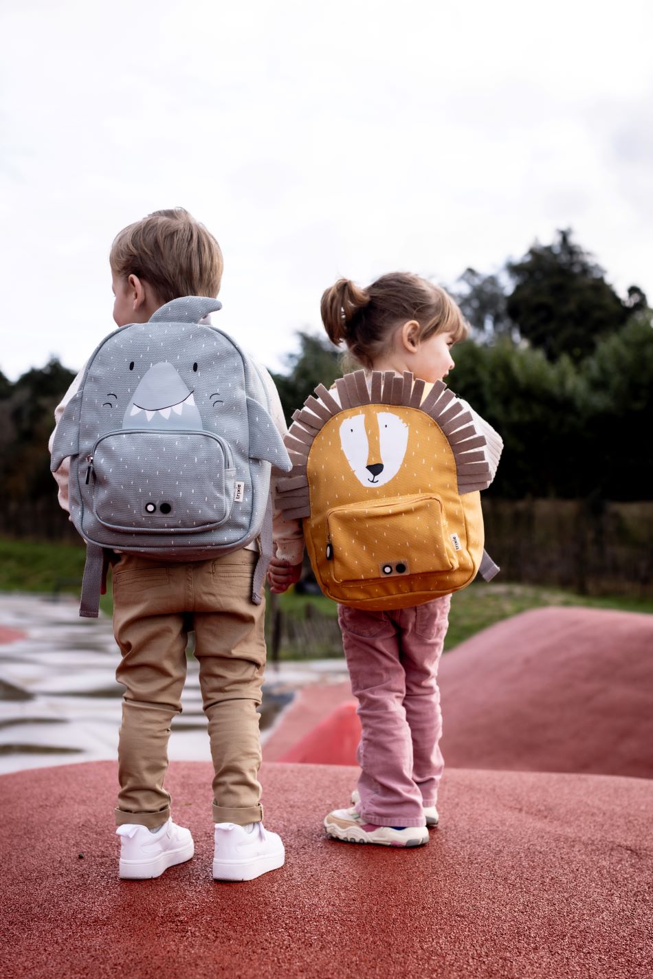 Duas crianças, um rapaz com a mochila Trixie Tubarão e uma rapariga com a mochila Trixie Leão, de costas, a observar uma paisagem exterior.