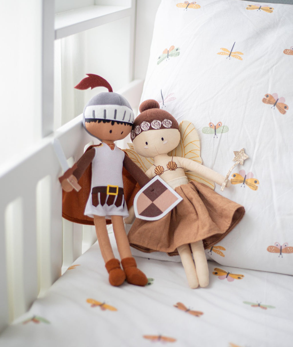 Peluches Cavaleiro Kai e Fada lado a lado num berço com roupa de cama estampada com borboletas.
