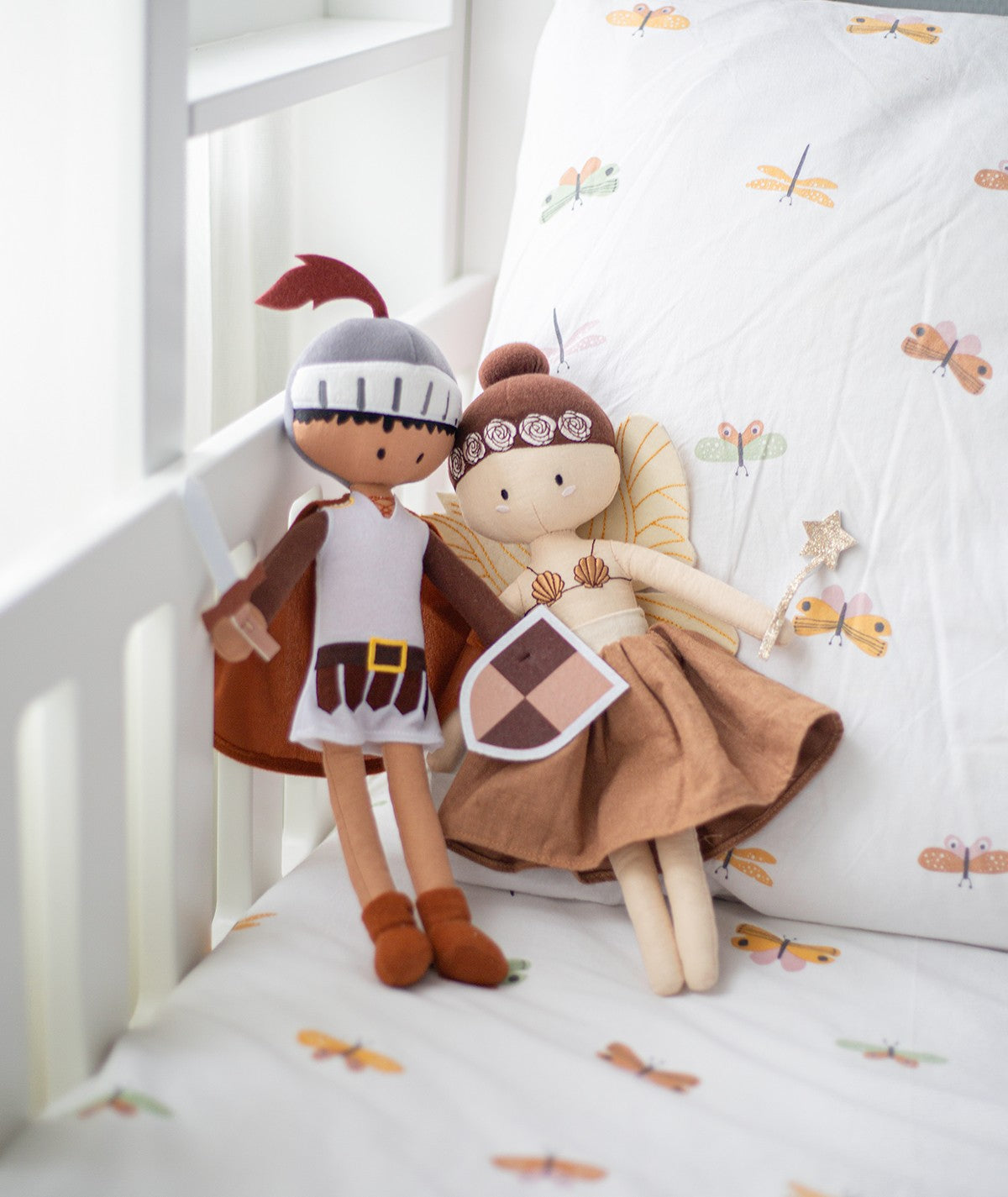 Peluches Cavaleiro Kai e Fada lado a lado num berço com roupa de cama estampada com borboletas.
